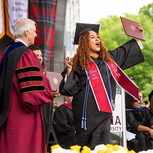A student holds her diploma and cheers on stage.