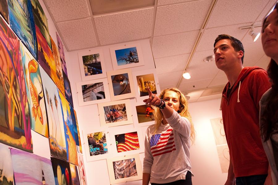 A student points at a wall of paintings in the Brother Kenneth Chapman Art Gallery as two other students look on.