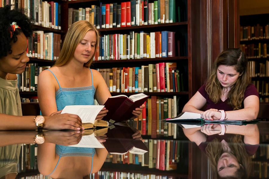 Three students sit at a table in the library reading books.