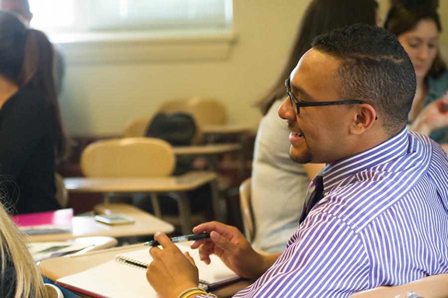A student with glasses and a beard in a shirt and tie smiles in a psychology class.