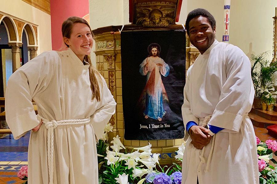 Two students where white robes to assist with Catholic mass with a painting of Jesus in between them.