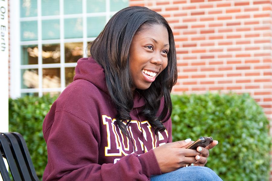A student sits on a bench and smiles and scrolls through her phone.