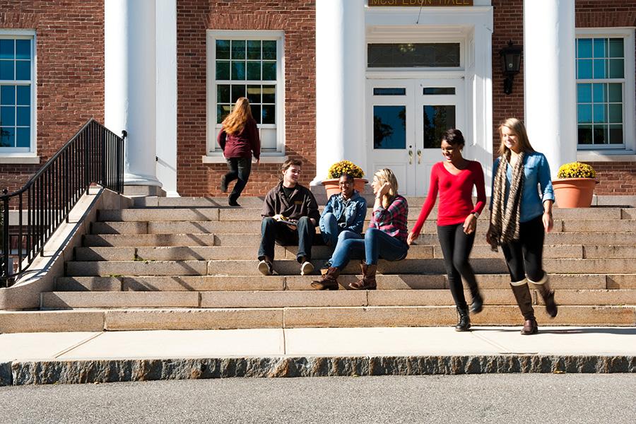 Three students sit on the steps while two students walk by and one student walks into the building.