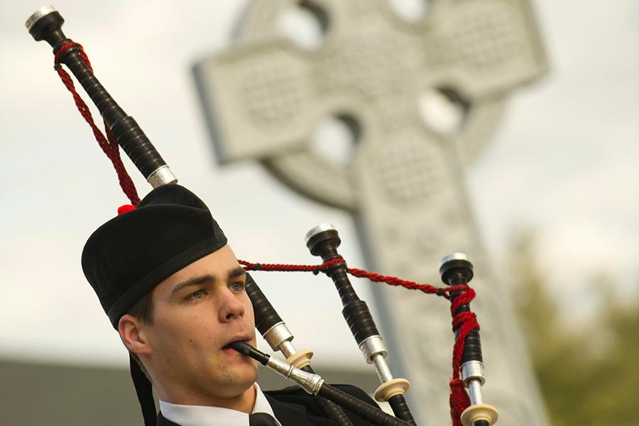 A piper plays bagpipes with the Celtic cross in the background.