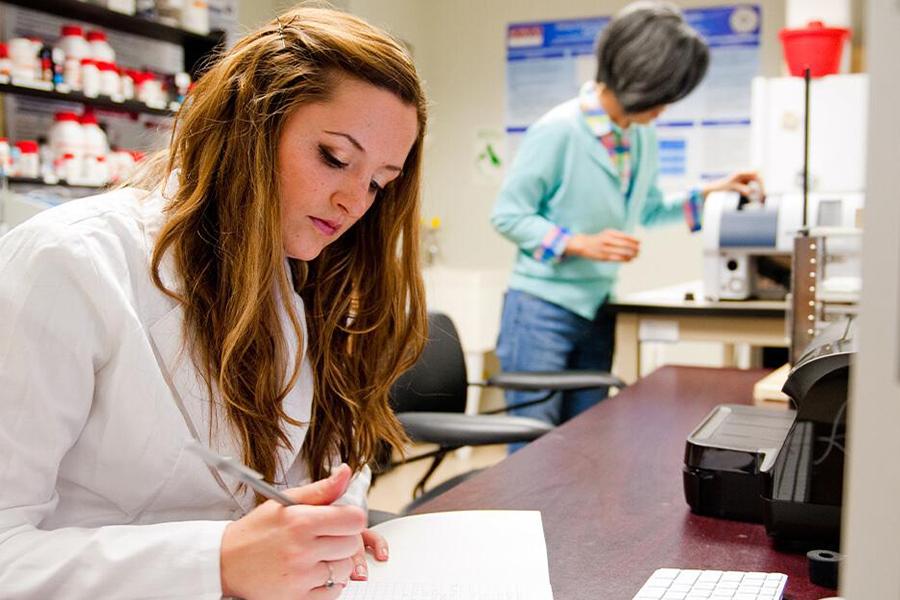 A student in a white lab coat writes something down while Sunghee Lee works in the background.