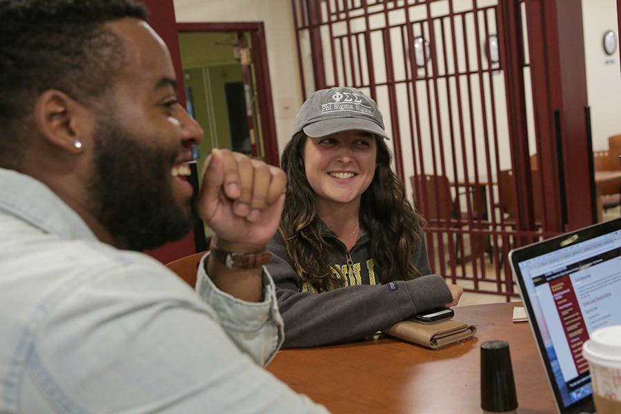 A student with a sorority cap smiles and talks to a student with a beard who smiles and laughs.
