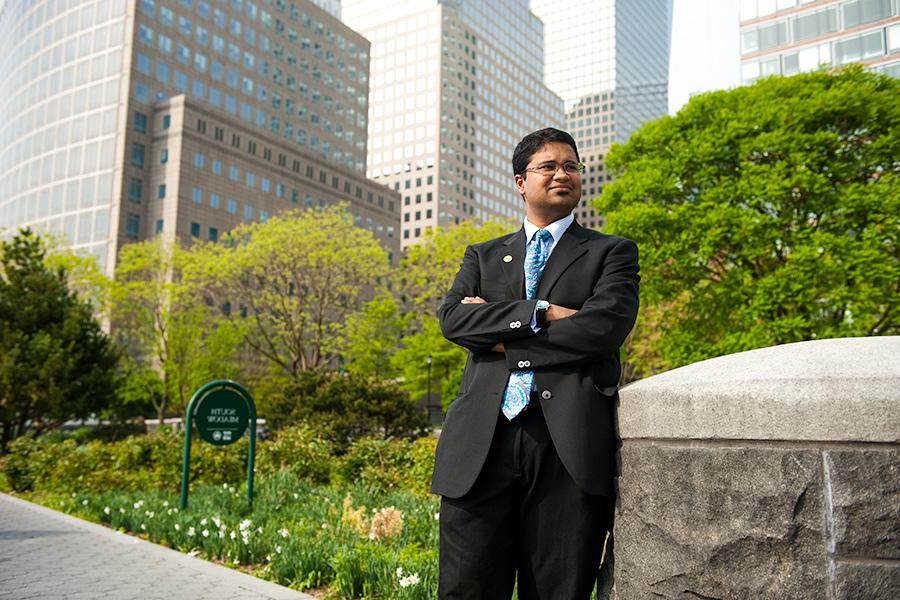 A student in a suit stands confidently in a park in New York City with his arms crossed.