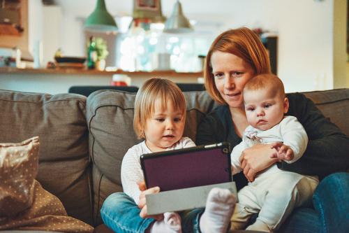 A mother looks at a digital tablet while her two children sit in her lap.