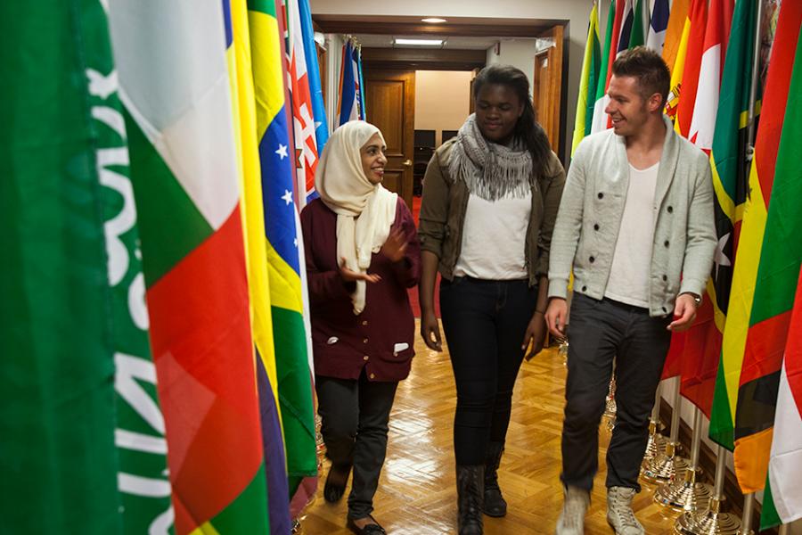 Three international studies students walks down a hallway lined with flags from different countries.