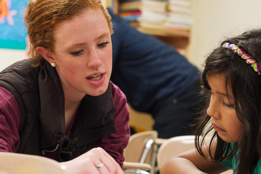 A female student sits next to a young girl, pointing to something on the desk top.