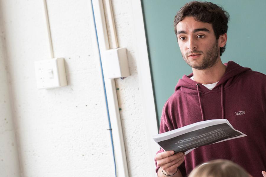 A male student stands reading a book in front of a class