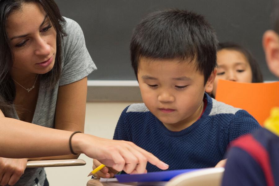 A female student sits beside a male elementary-age student pointing to a paper on the desk