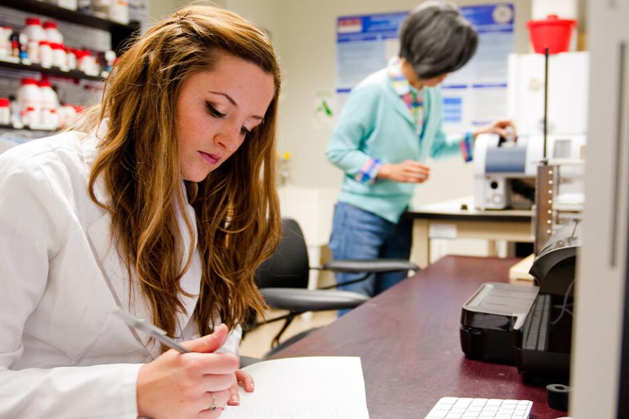 A female student in a lab coat works at a table in a lab with a female professor in background.