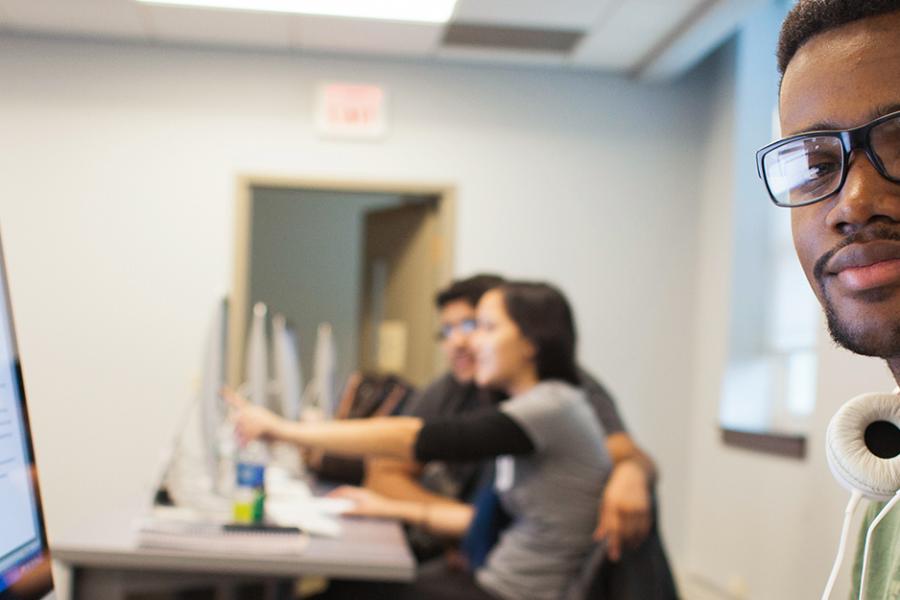 A student with white headphones around his neck works at a computer but looks toward the camera.