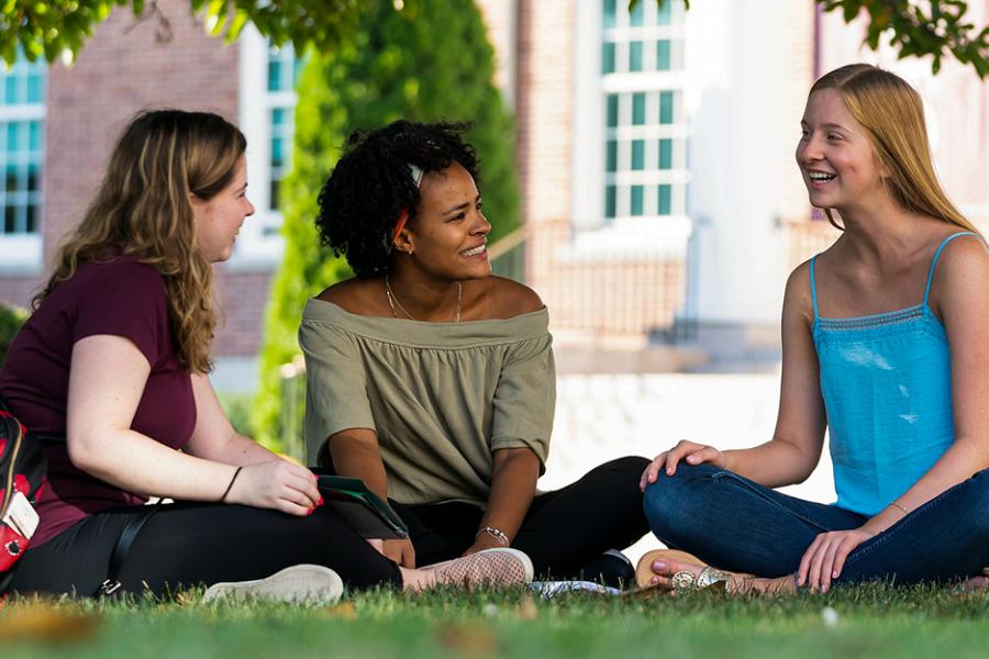 Three students sit under a tree on the grass and have a philosophical discussion.