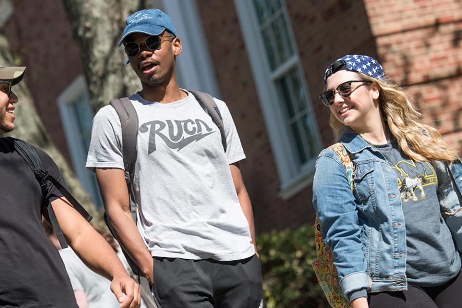 Two male students and one female student walk on campus and smile and talk. They are all stylish and artsy.