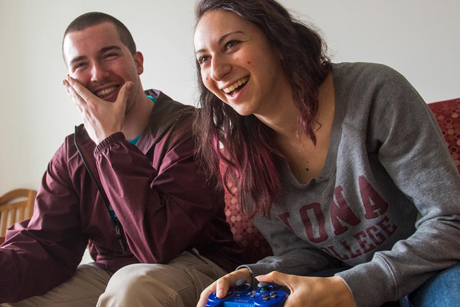 Two students play a video game together on a couch in a dorm room.
