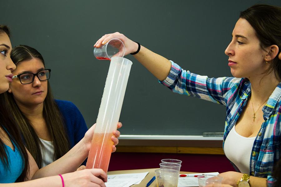 A group of chemistry students pour liquid from a beaker into a large graduated cylinder.