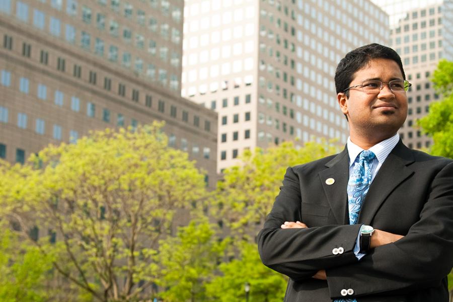 A student in a suit stands confidently in a park in New York City with his arms crossed.