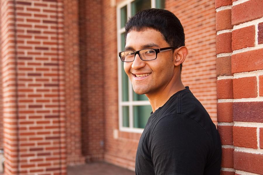 A student with glasses and a black tee shirt smiles and faces the camera outside of the Hynes Athletic center.