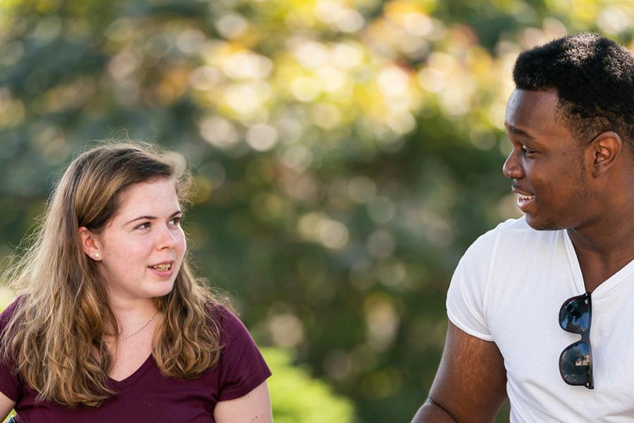 Two students sit outside and talk and smile on a sunny day.