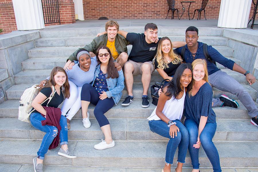 Iona students sit on the steps of Spellman and smile and give each other one arm hugs around the shoulders.