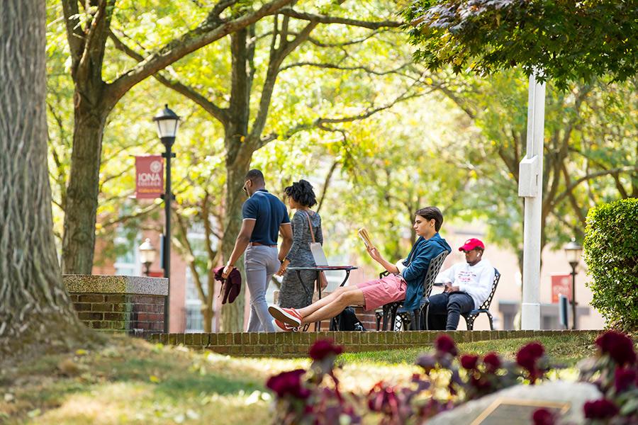 One Iona student sits outside and kicks his legs out and reads a book. Another sits at a different table working on homework. A man and woman hold hands and walk by.