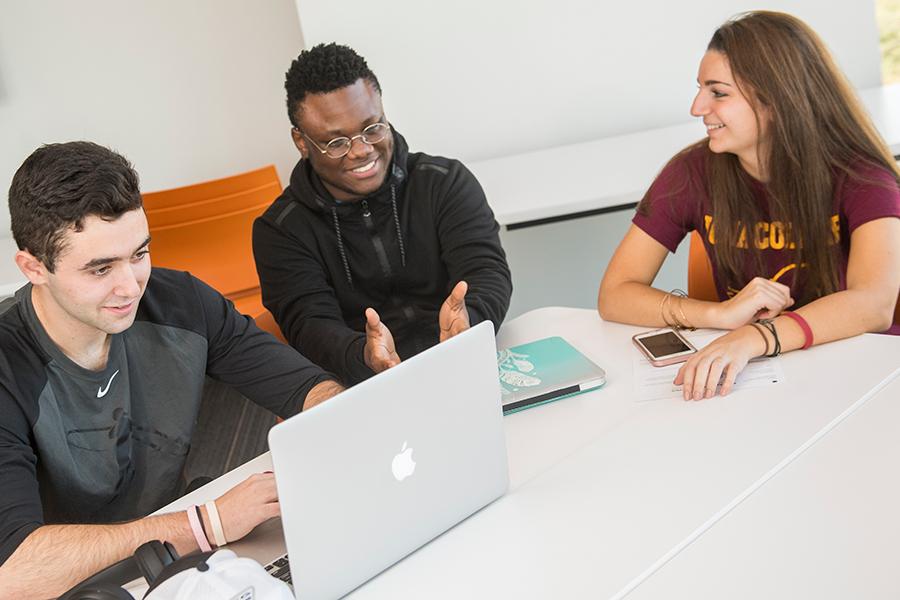 Three Iona students sit at a table in the North Ave. building with a mac laptop and one student is pointing something out.