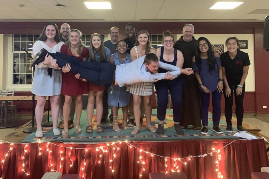 The Office of Mission and Ministry on a stage in the LaPenta Student Union. The first row of students hold another student as he lays across smiling.