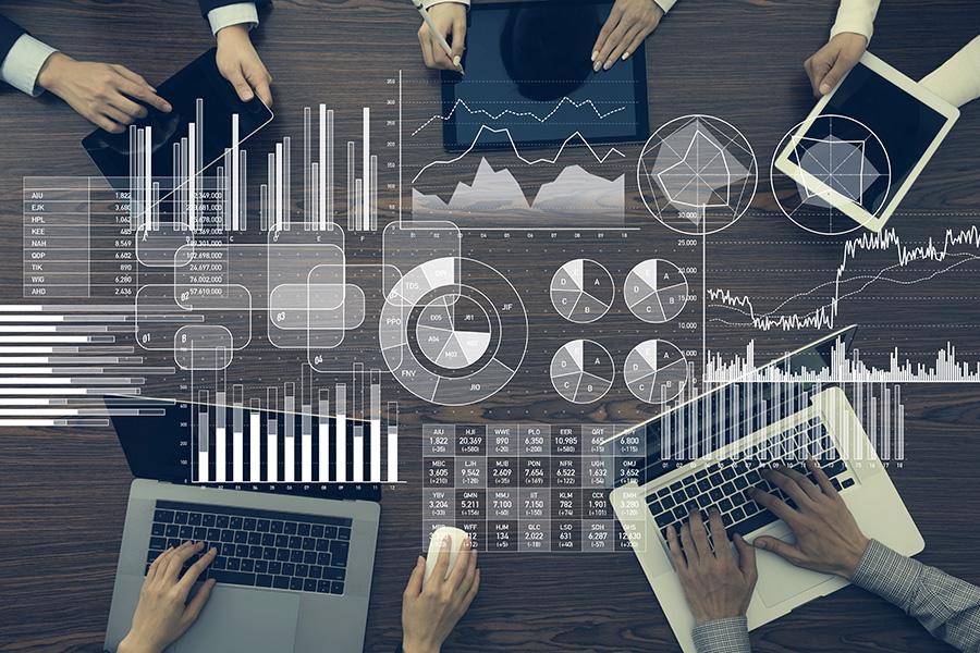 Overhead view of 5 business people at table, each working on an electronic device. Graphics of graphs and business numbers have been superimposed over the image.