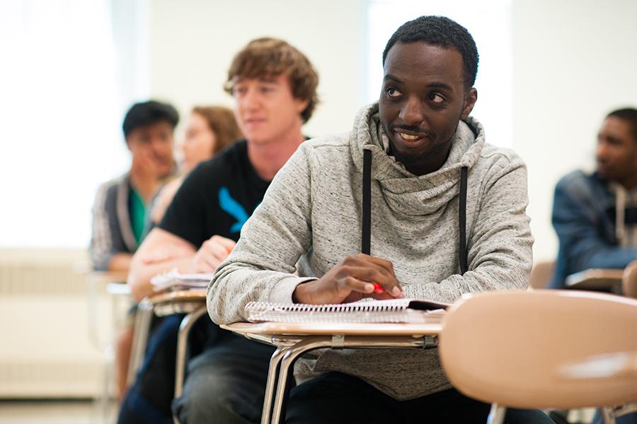A student smiles in class. He is in the forefront and three other students are in the row behind him.