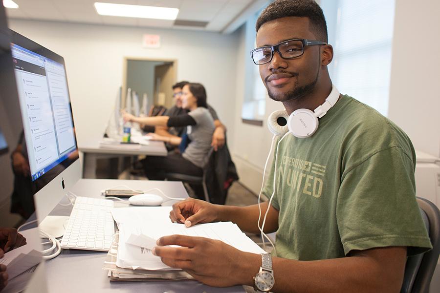 A student with white headphones around his neck works at a computer but looks toward the camera.