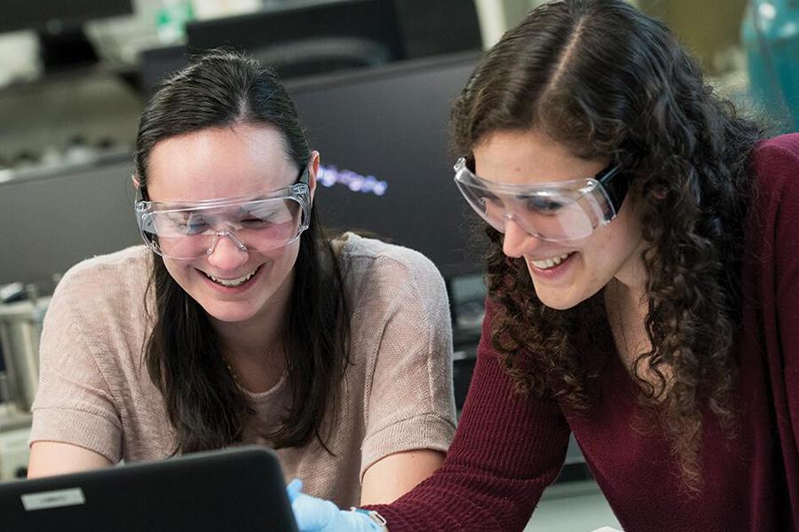 Two biology students wear goggles while reading something from a computer during an experiment.