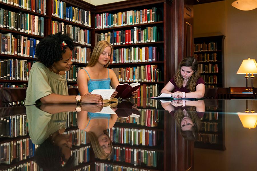Three students sit at a table in the library reading books.