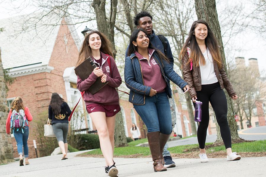 A group of three females and one male student walk on the pathway outside Amend Hall