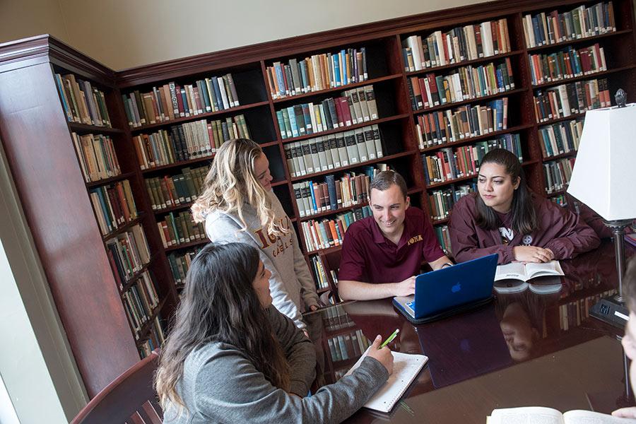 Four students sit at a table in the library, one works on a laptop and one writes in a notebook.