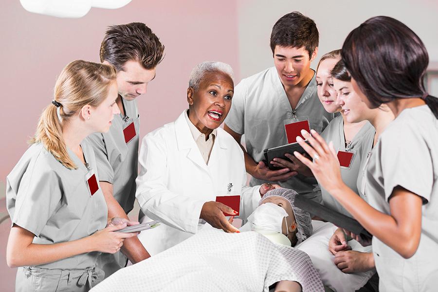 A female nursing instructor speaks to a mixed group of students in scrubs while pointing to a mannequin