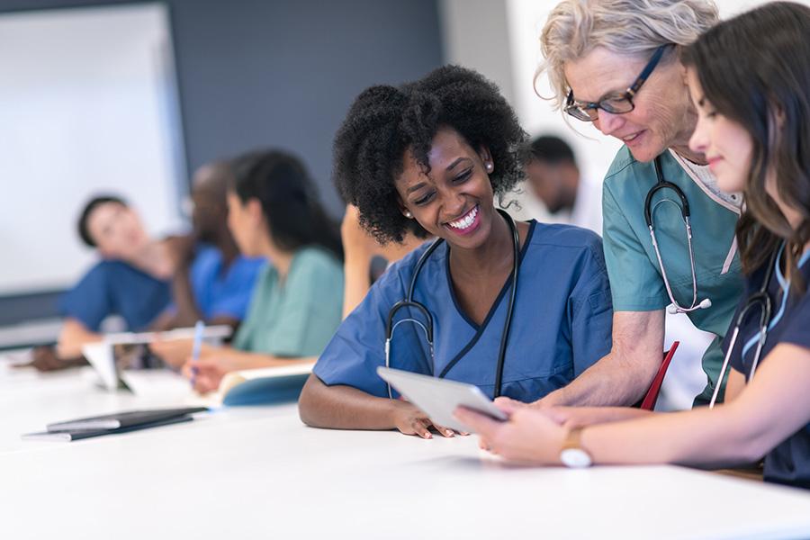Female nurses wearing scrubs sit at a table in discussion with other nurses in the background.
