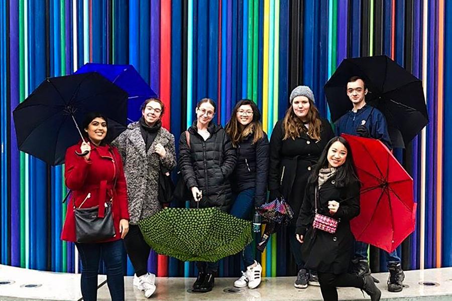 A group of Iona students pose in front of a rainbow colored wall while some hold umbrellas.