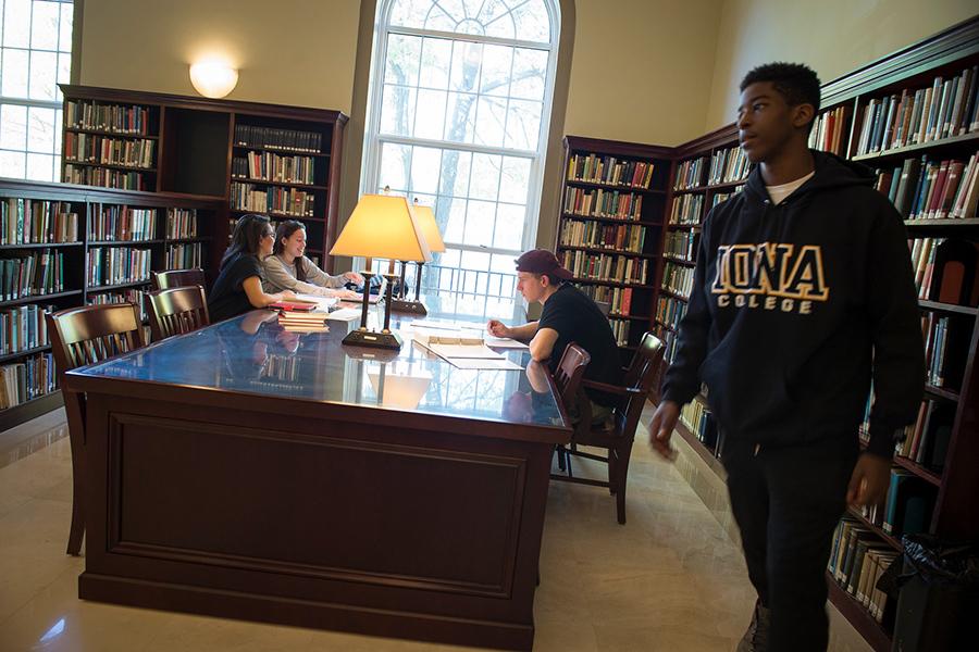 Two female students work at a table in Ryan Library and smile. One male student works across from them and another male student in a Iona sweatshirt walks past.