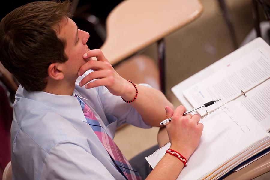 A student in a tie and button up shirt with his hand on his chin takes notes in a finance class.
