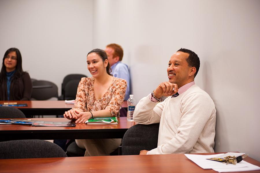 A student in a white sweater with a tie on leans on his hand and smiles during class.
