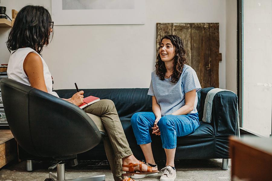 A therapist sits on a chair and talks to her patient on a couch.