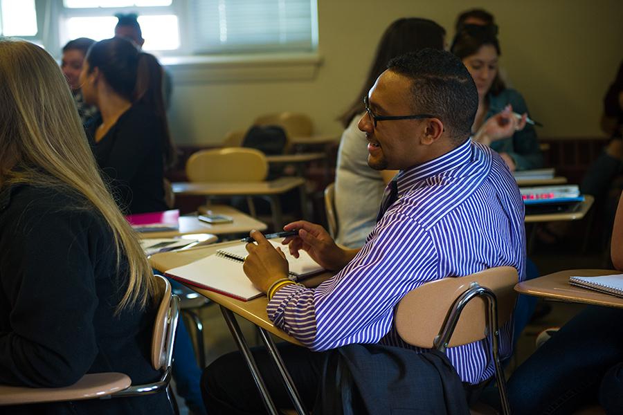 A student with glasses and a beard in a shirt and tie smiles in a psychology class.