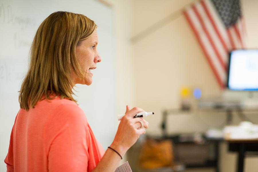 A professor lectures in class and holds a dry erase marker in her hand.