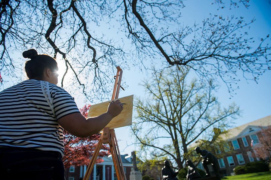 A female student stands at an easel painting in the campus quad.