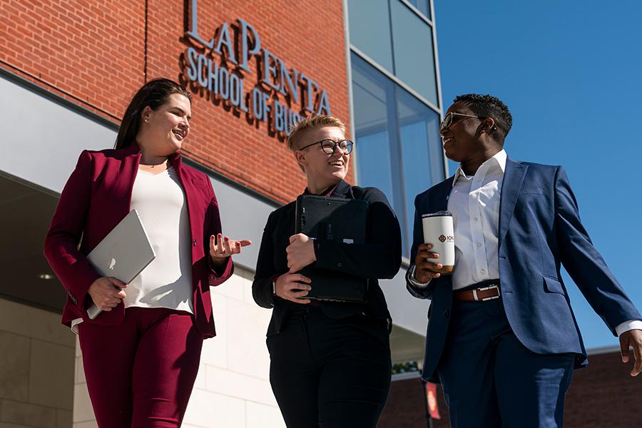 Three graduate business students walk in front of the LaPenta School of Business building on a sunny day.