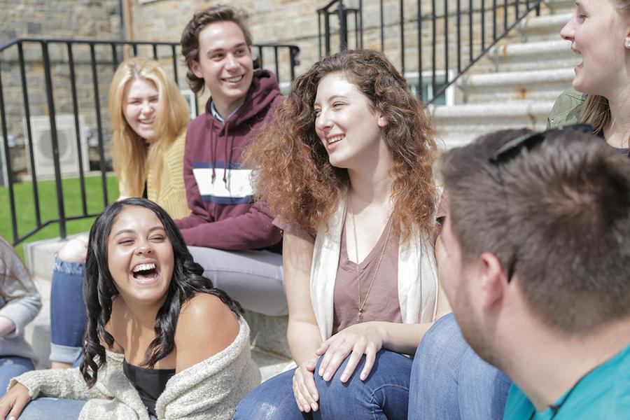 Students sit on the steps of Doorley Hall. A female student with black hair smiles widely and laughs.