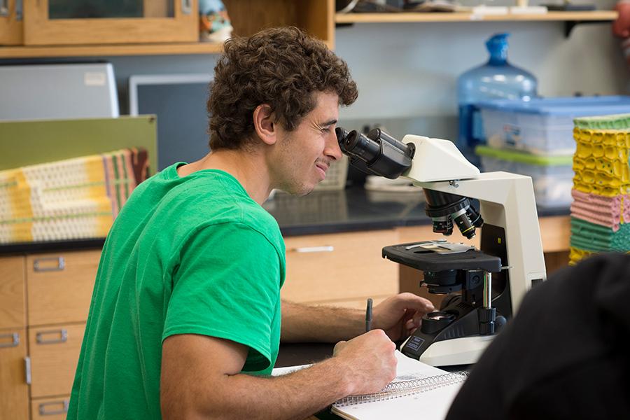 A student in a green shirt looks through a microscope in biology class.
