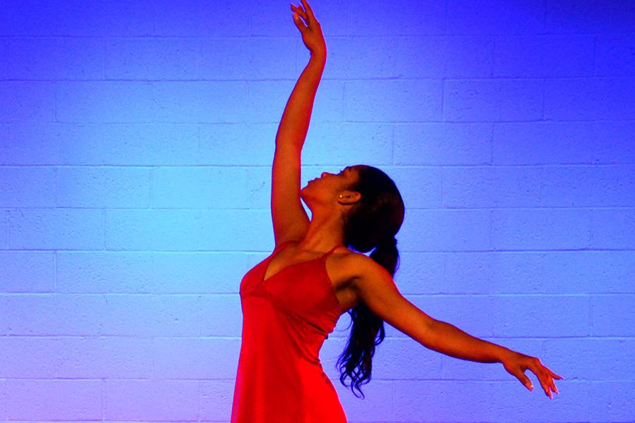 A dancer in a red dress raises her right arm and lowers her left arm.